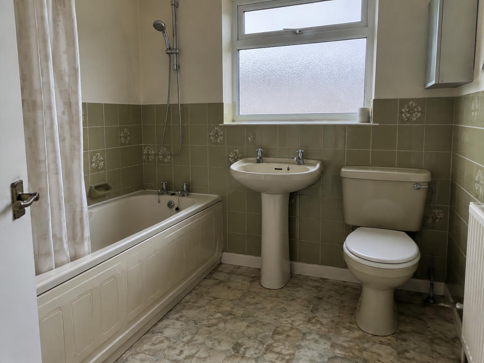 The same bathroom before renovation, with dated green tiling, a panelled bath and worn vinyl flooring